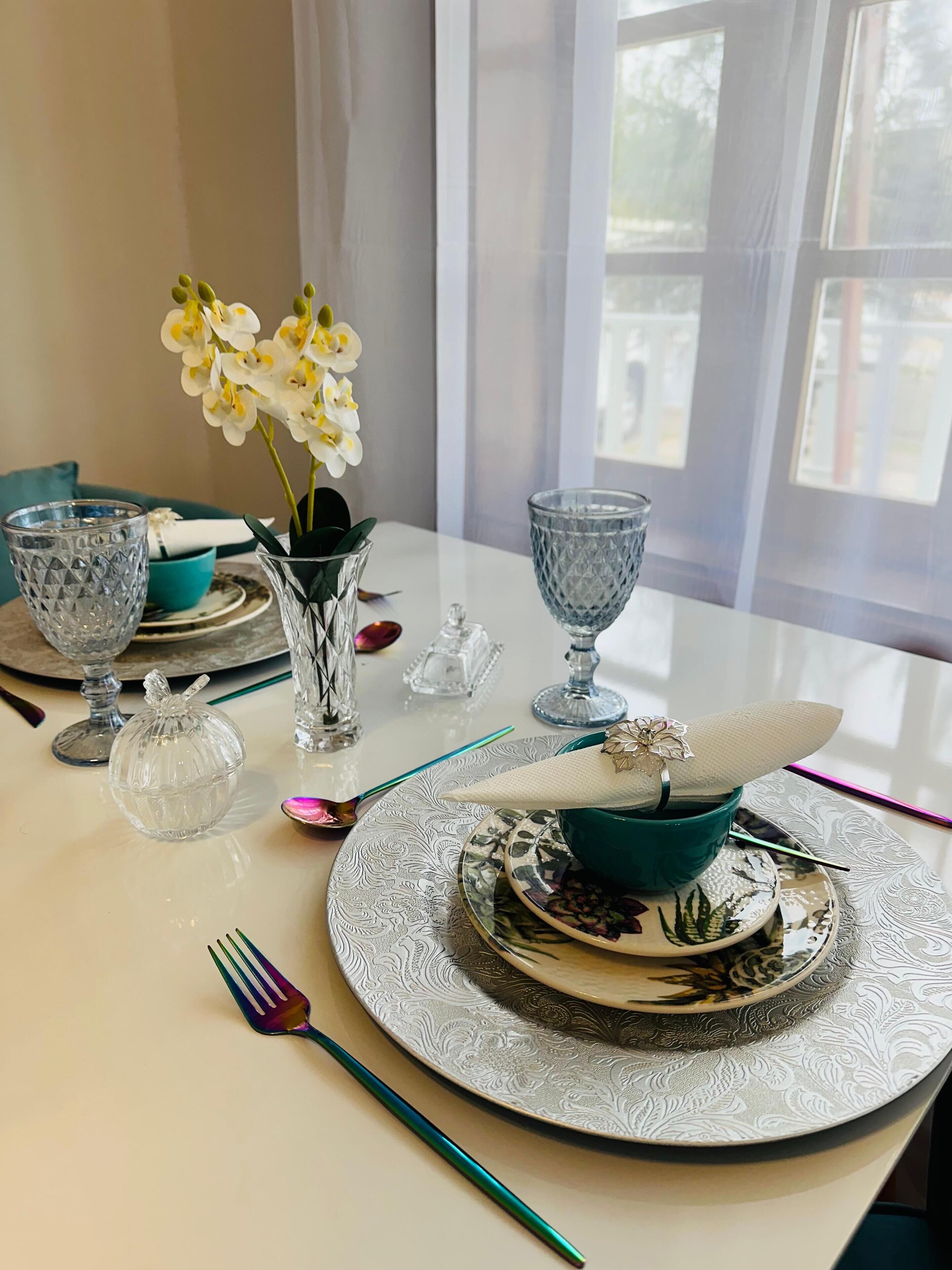 Ein Esstisch, gedeckt mit einer blauen Schale, einem klaren Glas, einer Kristallvase mit Blumen und einem dekorativen Teller.