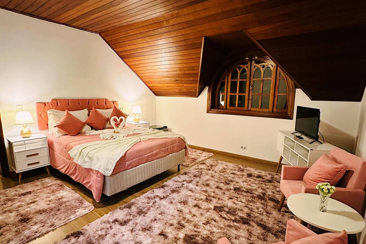 Quarto com teto inclinado, cama com roupa de cama rosa, tapete felpudo, poltrona rosa, mesinha branca pequena e janela com moldura de madeira.