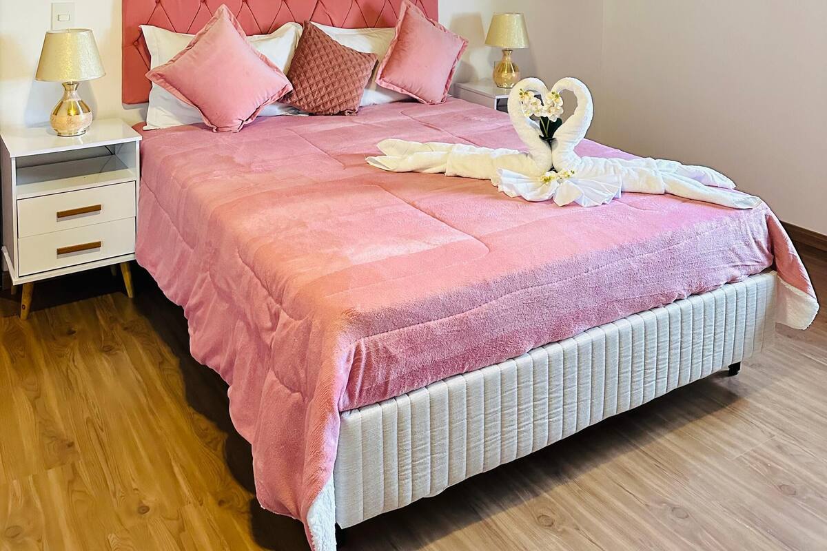 Quarto com teto inclinado, colcha rosa e almofadas combinando. Há duas mesas de cabeceira com abajures.