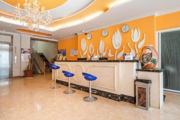 Zona de recepción de hotel con mostrador, taburetes de bar, una araña y una pared con detalles decorativos.
