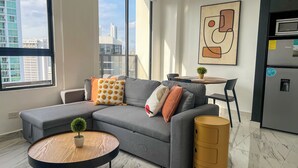 A modern living room with a grey sectional sofa, a small wooden coffee table, a potted plant, and a compact kitchen area with a refrigerator and a sink.