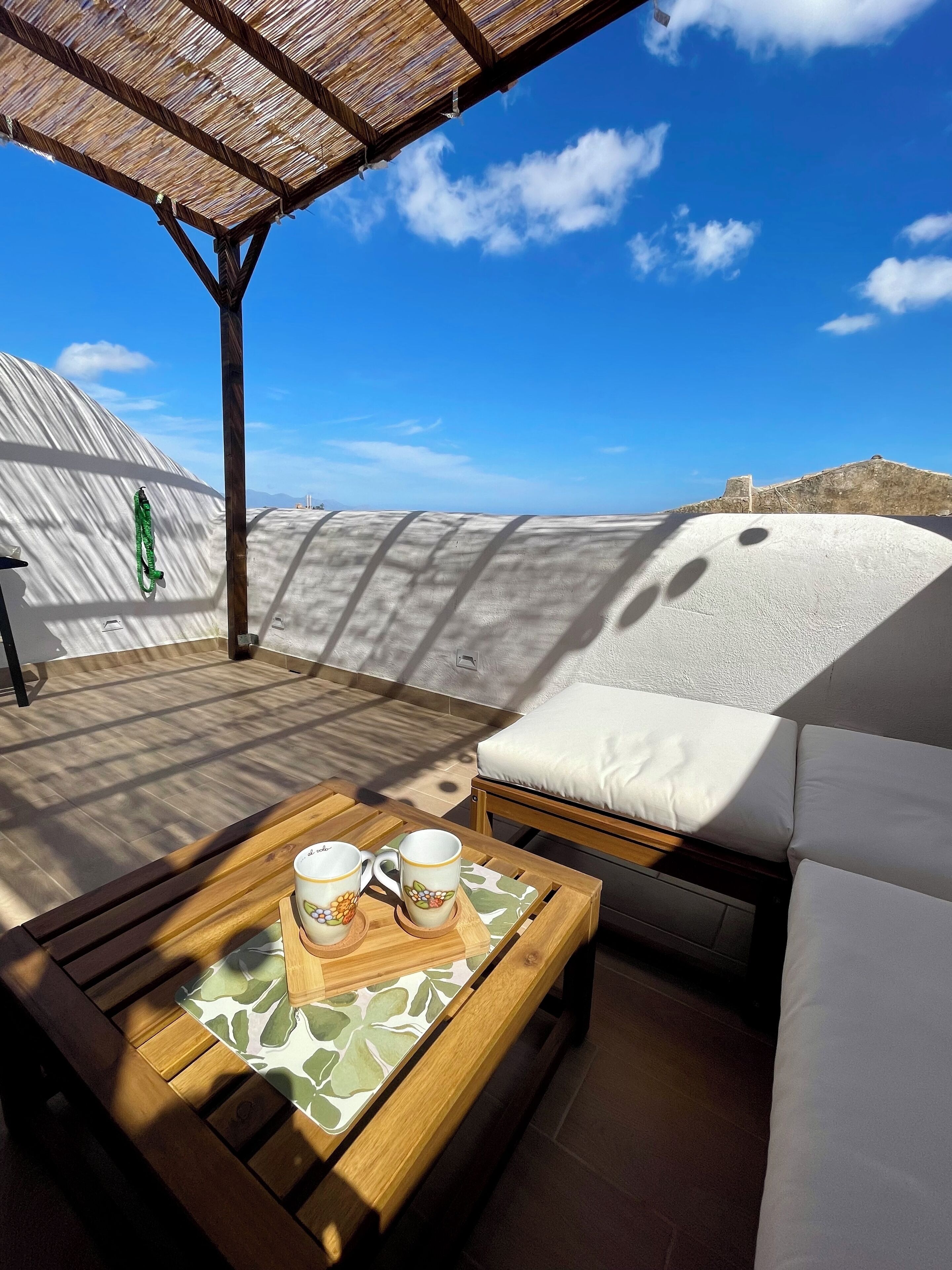 Un espace extérieur ensoleillé, aménagé avec une table en bois, un canapé blanc doté de coussins et une pergola au-dessus.
