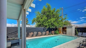 Une piscine rectangulaire à l’eau d’un bleu limpide, entourée d’une terrasse en béton. On y trouve des chaises longues et une petite table avec des coussins. Un grand arbre aux feuilles vertes et luxuriantes est visible à l’arrière-plan, et un mur de briques orné d’un motif décoratif se trouve derrière la piscine.