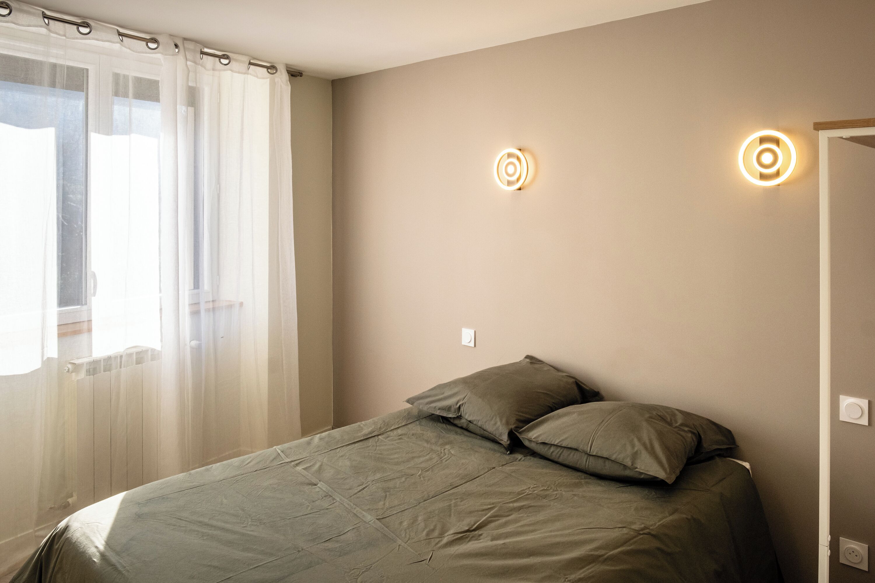 Un dormitorio con una cama, un ventanal con cortinas traslúcidas y dos lámparas de pared.