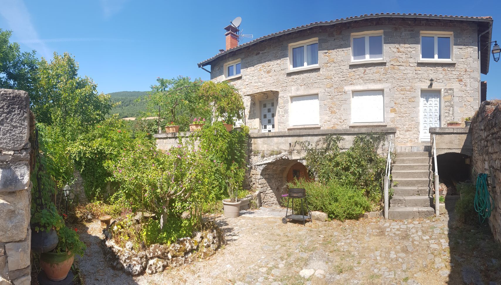 Una casa de piedra con una escalera de piedra que conduce a una puerta, rodeada de follaje y un cielo despejado.