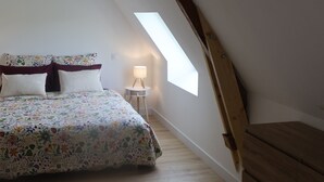 A bedroom with a sloped ceiling, a bed with floral bedding, a bedside table with a lamp, and a wooden dresser. - l'épi dort (Saint Maclou la Briere)
