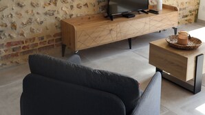 A modern living room with a brick-patterned wall, a wooden console table with a flat-screen TV, a grey sofa, and a coffee table with a woven basket. - l'épi dort (Saint Maclou la Briere)