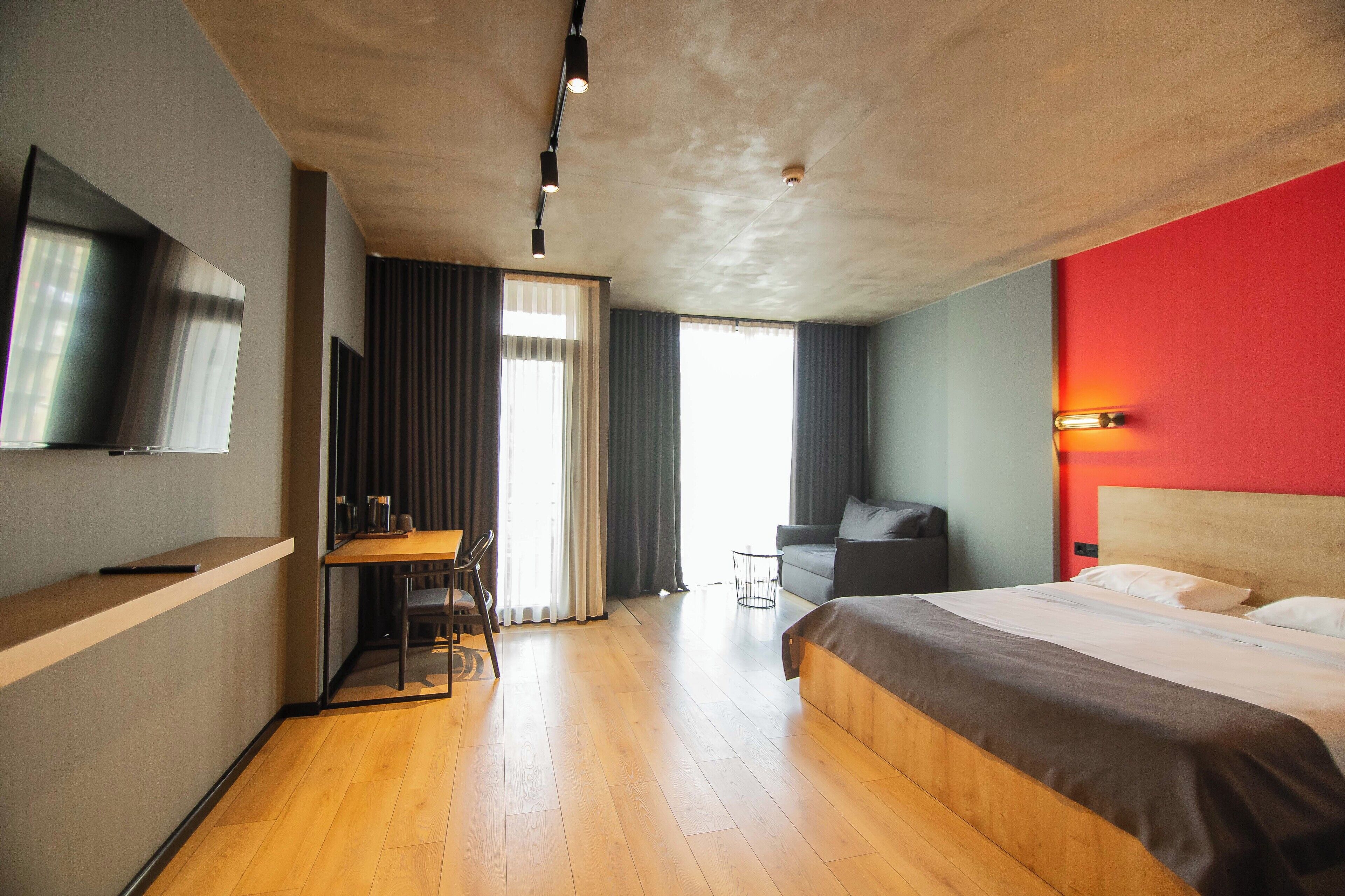 Een hotelkamer met een bed, een bureau met stoel, een aan de muur bevestigde televisie en een groot raam met gordijnen.