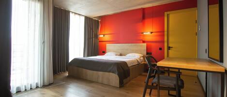 A modern hotel room with a wooden floor, a bed with a headboard, a desk with a chair, and a large window with curtains.