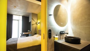 Camera d'albergo moderna con letto, scrivania e bagno dotato di un ampio specchio e lavandino.