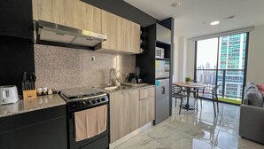 Cocina moderna con estufa a gas, pileta y horno empotrado. Hay una zona de comedor con mesa y sillas, y un ventanal con vista a la ciudad.
