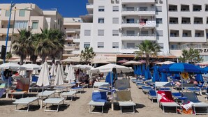 Una playa con filas de tumbonas y sombrillas, un edificio de varios pisos con balcones, y un letrero que dice "Blue Royal Apartment".
