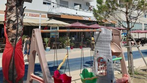 Un parque infantil con columpios y un tobogán, palmeras y un edificio de varios pisos con balcones al fondo.