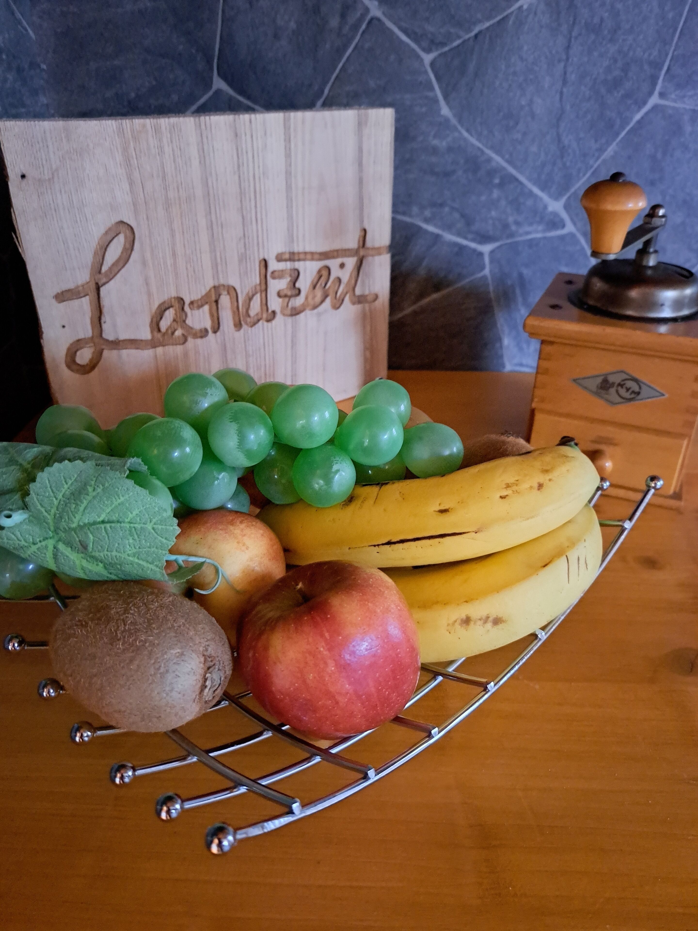 Eine Obstschale mit Trauben, Bananen, einem Apfel und einem Kiwifrucht auf einem Drahtgestell.