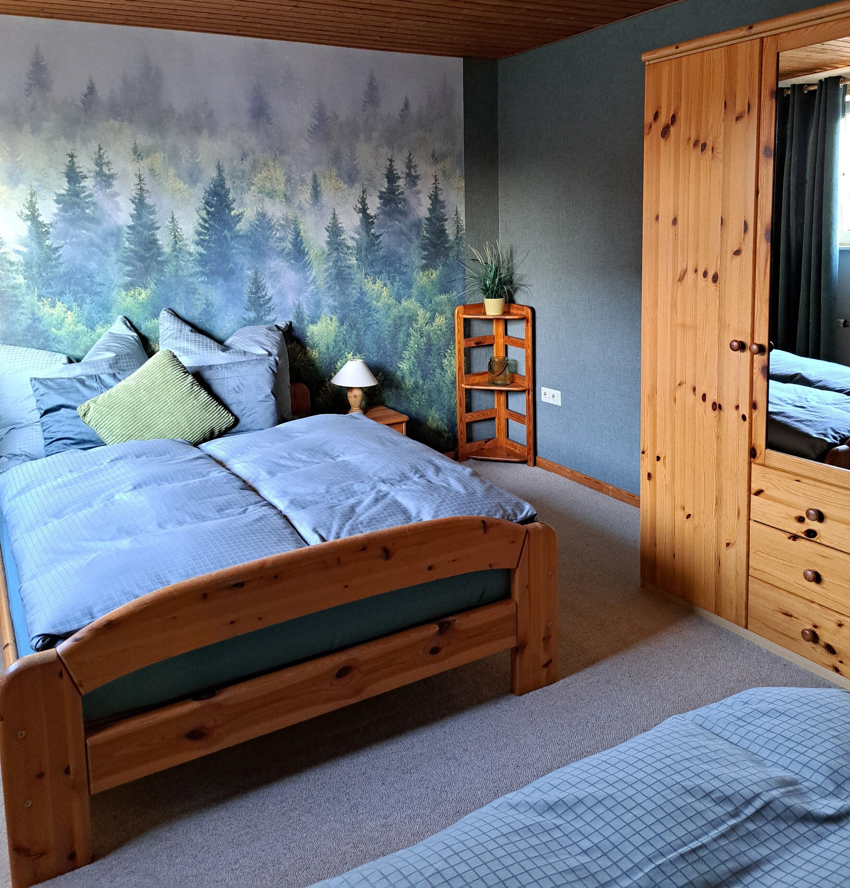 Une chambre à coucher comprenant un lit, une armoire en bois, une table de chevet avec une lampe et une fresque murale représentant une forêt.