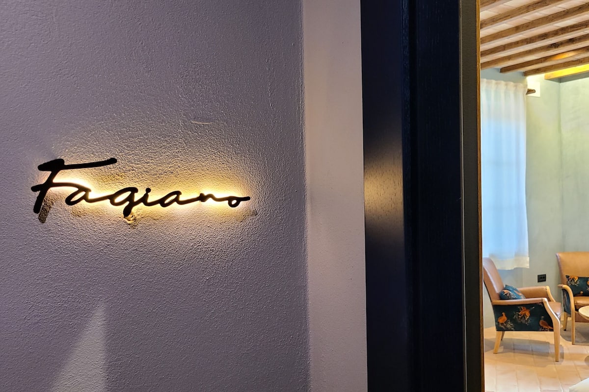 Um quarto com uma placa na parede com a inscrição 'Fagiano', teto de madeira, uma pequena mesa redonda, duas cadeiras e uma almofada estampada.