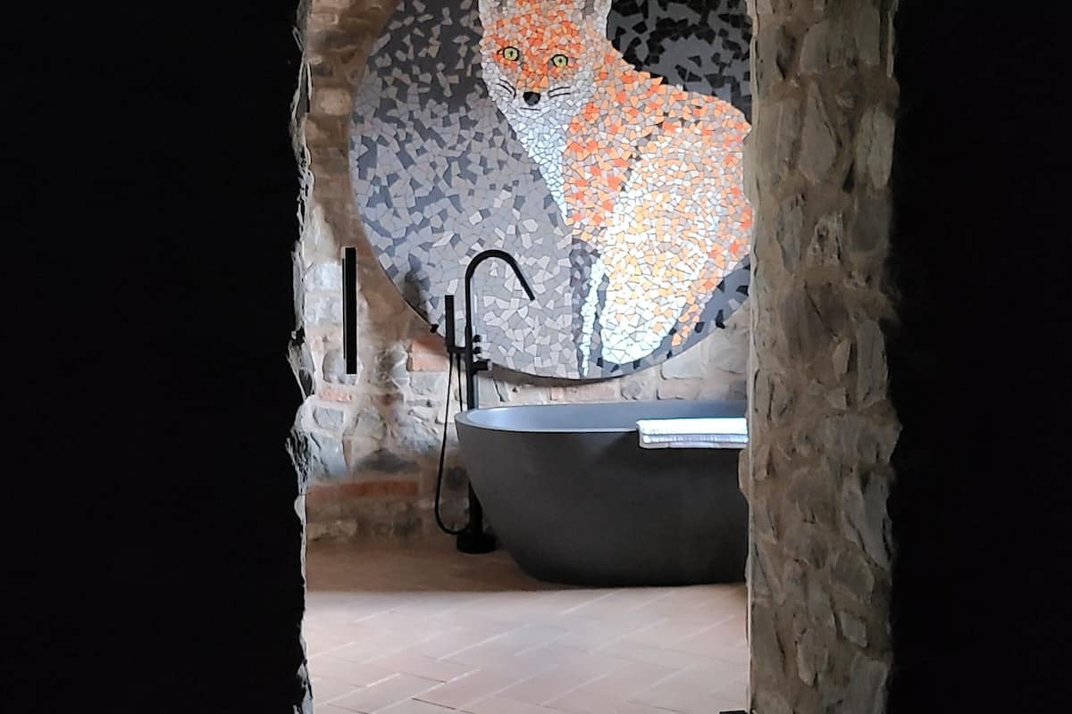 Casa de banho com paredes de pedra, um mosaico de uma raposa, uma banheira e um chuveiro.