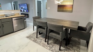 A modern kitchen with a built-in oven, a dining area with a wooden table and grey chairs, and a TV mounted on the wall.