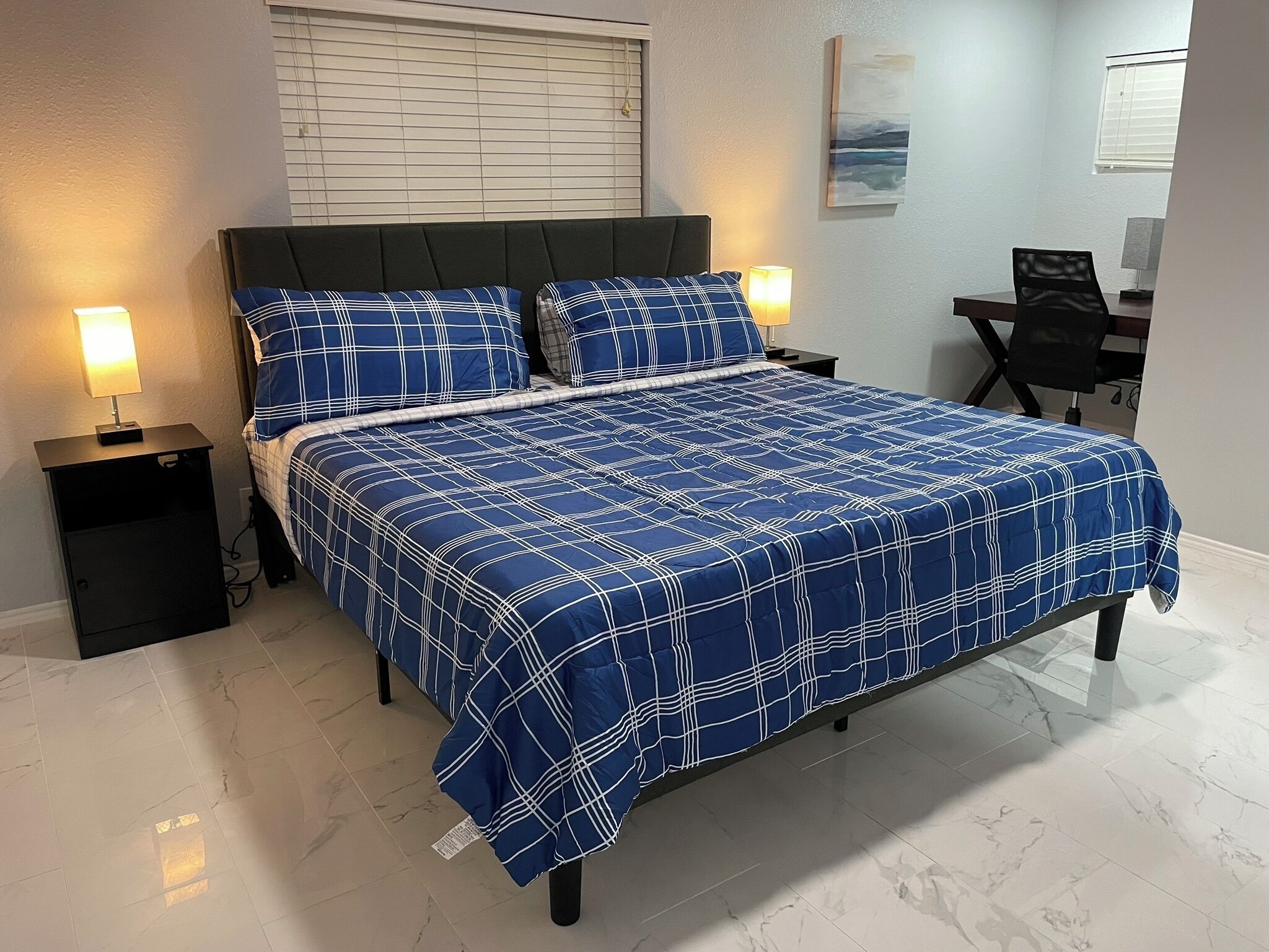 A neatly made bed with a blue and white checkered bedspread, flanked by two bedside tables with lamps, a black chair, and a desk with a lamp.