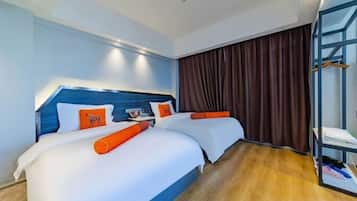 Habitación de hotel con dos camas, cabecero azul, ropa de cama blanca, almohadas naranjas, suelo de madera y un ventanal con cortinas.