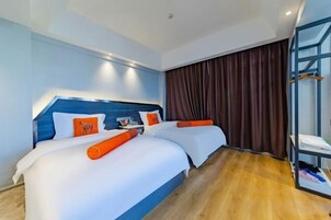 Habitación de hotel con dos camas, cabecero azul, ropa de cama blanca, almohadas naranjas, suelo de madera y un ventanal con cortinas.
