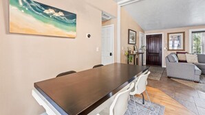 A dining area with a long table and chairs, a painting on the wall, and a living room with a sofa and a door in the background. - Charming Mountain Retreat with Enclosed Backyard and Artistic Flair (Millcreek)
