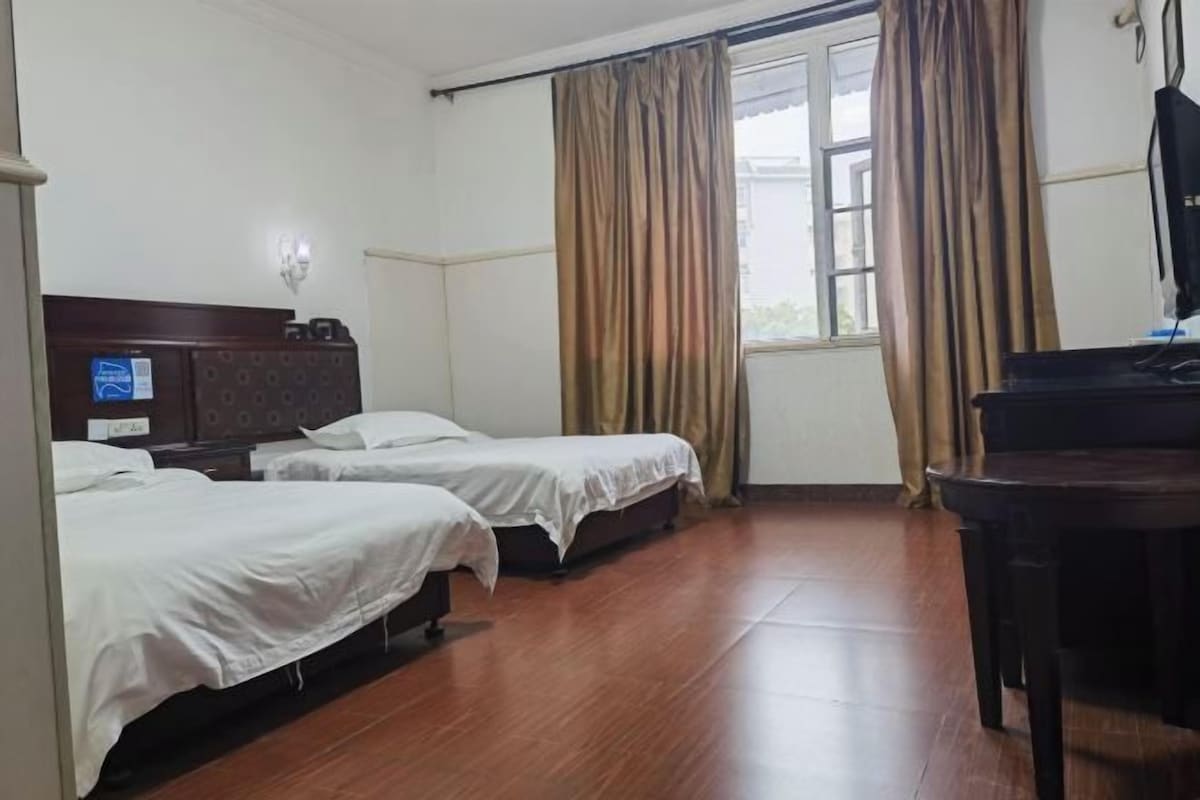 A hotel room with two single beds, a wooden floor, a desk with a chair, and a window with curtains.