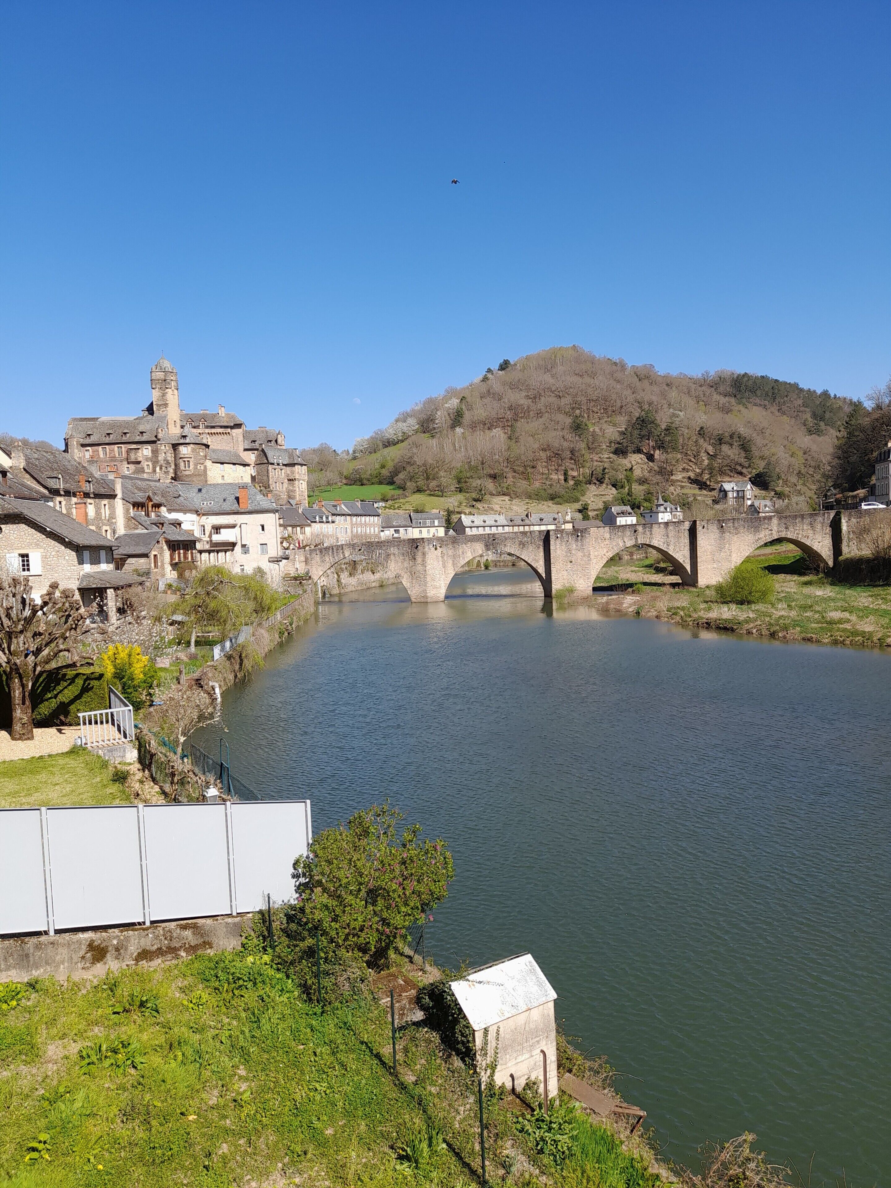 Un pont de pedra històric sobre un riu, una vila amb arquitectura tradicional i un turó al fons.