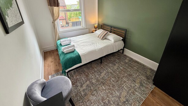 A small, neatly arranged bedroom with a single bed, a chair, a window with curtains, and a view of the outside. - The Walthamstow Lodge (London)