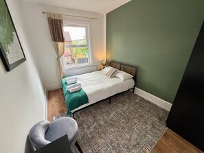 A small, neatly arranged bedroom with a single bed, a chair, a window with curtains, and a view of the outside.