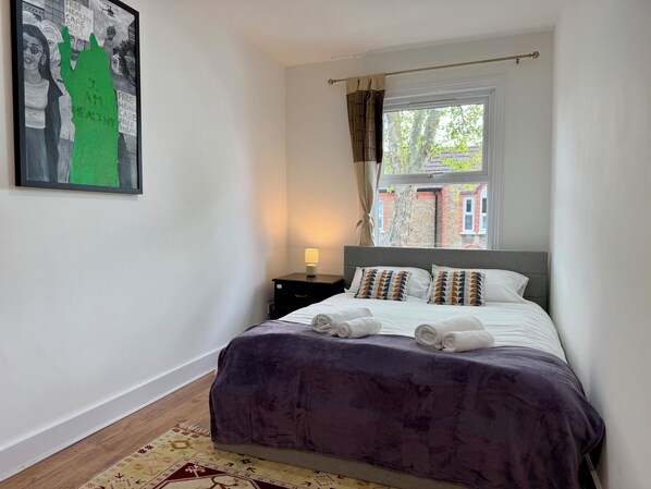 A bedroom with a bed, a nightstand, a window with curtains, and a painting on the wall.