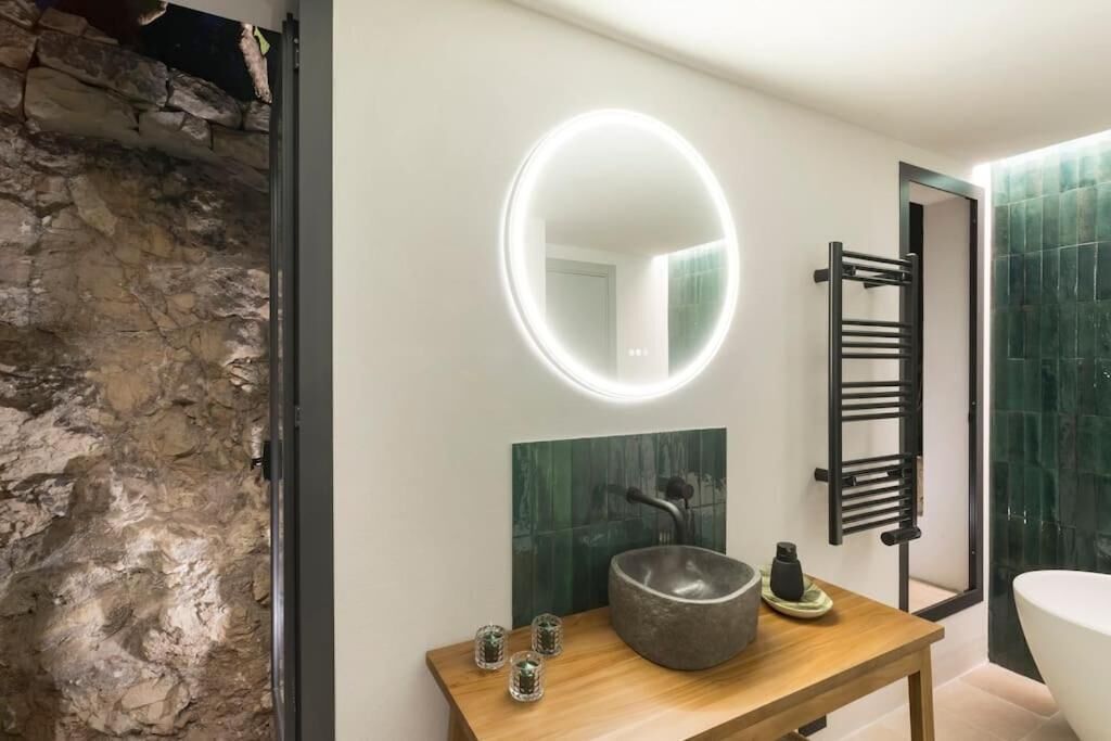 A modern bathroom with a stone wall, a round mirror with a light, a wooden vanity with a large basin, and a towel rack.