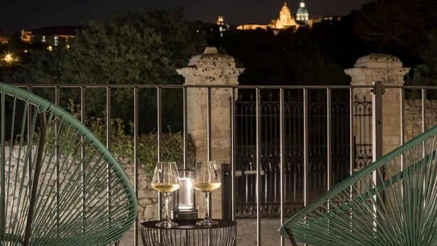 Un balcón con dos sillas verdes, una mesa redonda pequeña y tres copas de vino blanco. Al fondo, se ve un edificio con una cúpula y luces.