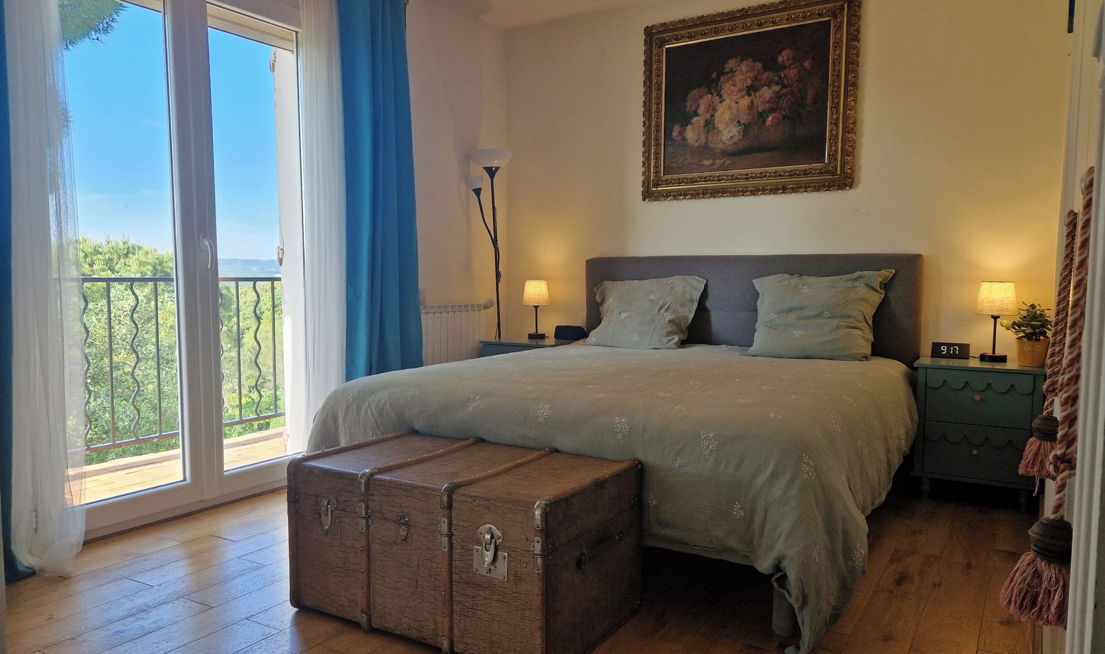 Une chambre à coucher avec un grand lit, une table de chevet, une lampe, un tableau, une valise et une vue par la fenêtre.