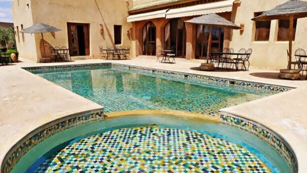 A swimming pool with a mosaic tile design, surrounded by outdoor seating and a building with a balcony. - Riad dar chenguiti (Taroudant)