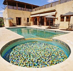 A swimming pool with a mosaic tile design, surrounded by outdoor seating and a building with a balcony. - Riad dar chenguiti (Taroudant)