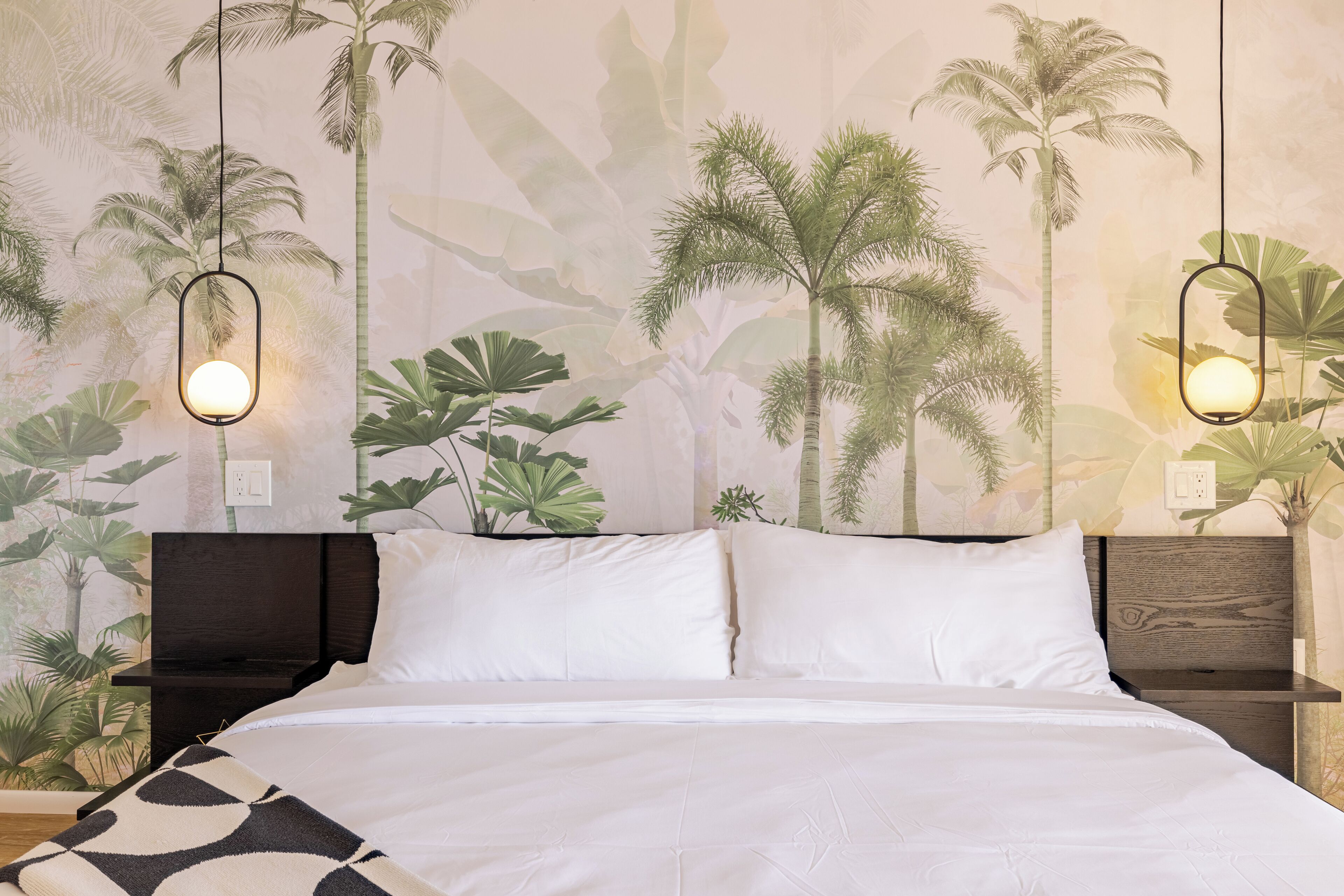 A bedroom with a bed, white bedding, two nightstands, and a wall mural of tropical plants.
