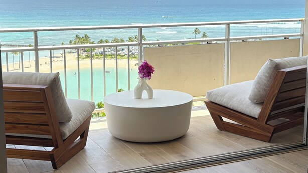 Een balkon met uitzicht op de oceaan, twee houten stoelen, een ronde witte tafel en een vaas met bloemen.