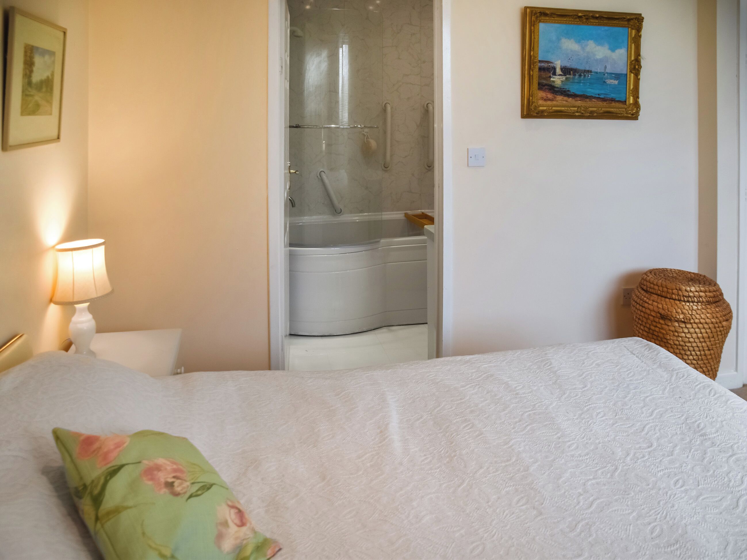 Une chambre à coucher avec un lit, une table de chevet, une lampe, un tableau, et une salle de bain visible à travers une porte ouverte.