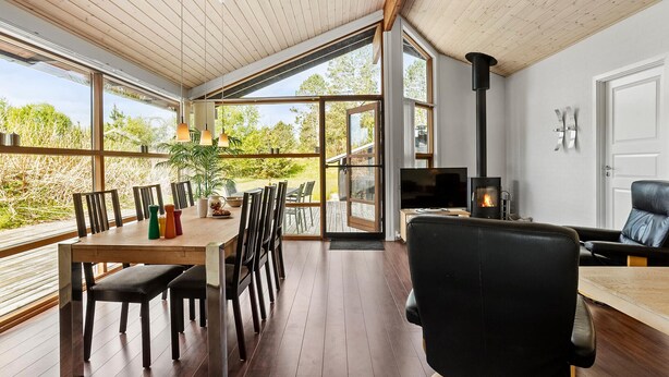 A spacious room with a wooden ceiling, large windows, a dining area with a table and chairs, a black leather sofa, and a fireplace. - Stunning home in ålbæk with sauna (Ålbæk)
