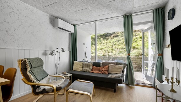Ein modernes Wohnzimmer mit einer Couch, zwei Sesseln, einem kleinen Tisch und einer Stehlampe. Es gibt ein großes Fenster mit Vorhängen sowie eine Klimaanlage an der Wand.