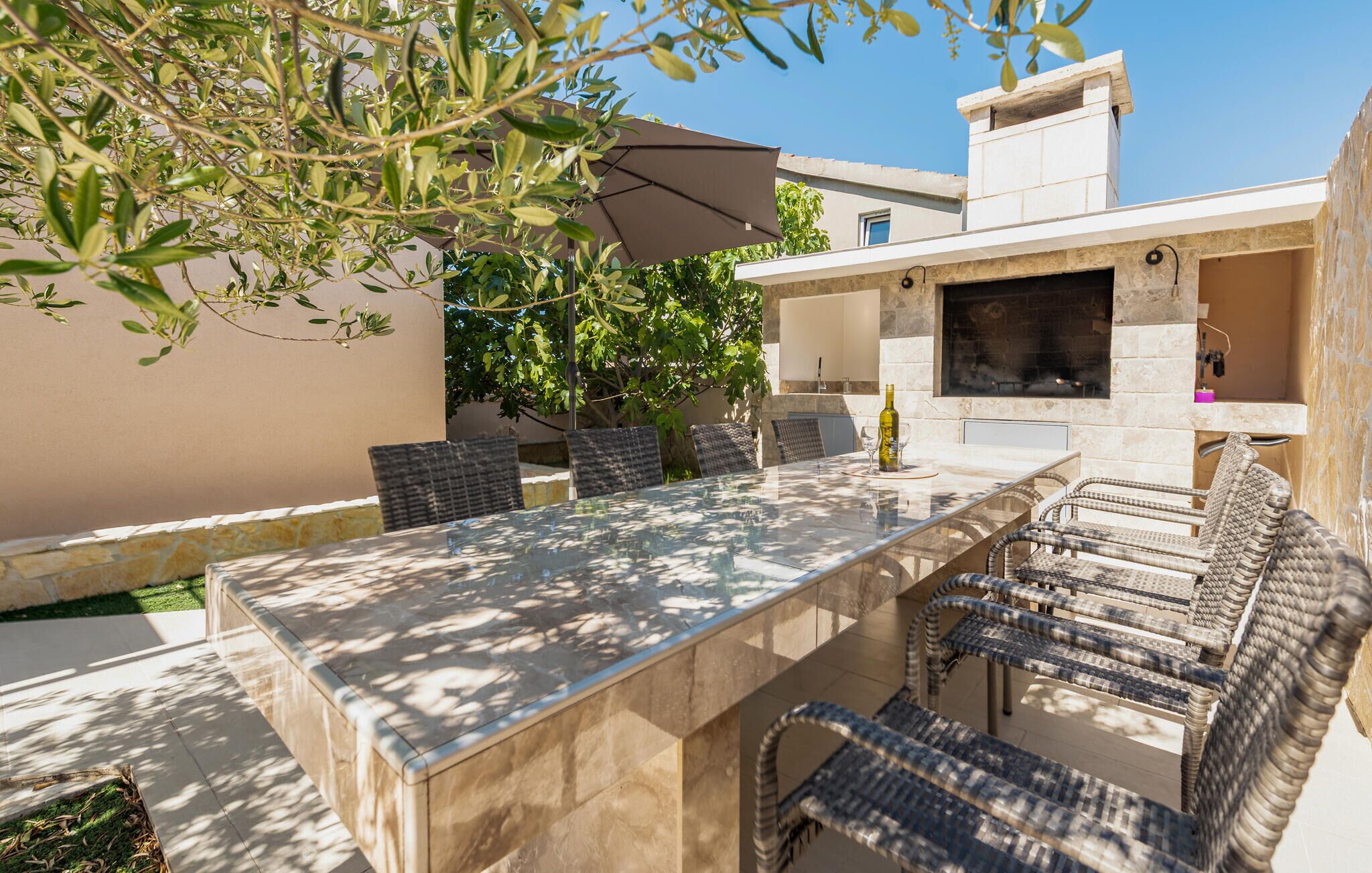 An outdoor dining area with a stone table and wicker chairs, a pergola with a brown umbrella, and a stone fireplace.