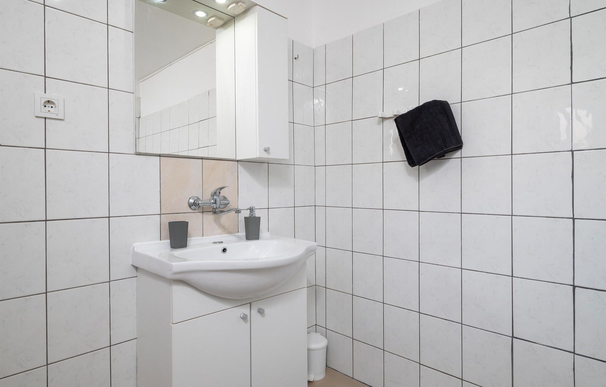 A bathroom with white tiled walls, a white sink with two faucets, a mirror above the sink, and a black towel hanging on the wall.