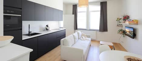 Et moderne køkken med mørke skabe, en hvid bordplade og en indbygget ovn. I opholdsstuen er der en hvid sofa og trægulv. Et spisebord med en skål nødder er synligt.