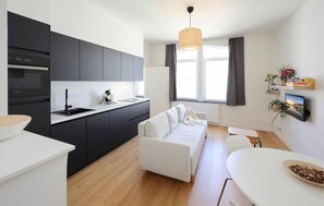 Une cuisine moderne avec des meubles sombres, un plan de travail blanc et un four encastré. Le salon est doté d’un canapé blanc et d’un parquet en bois. On peut apercevoir une table à manger avec un bol de noix.