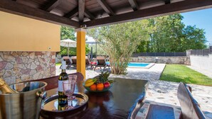 A covered outdoor area with a pool, lounge chairs, and a table with a bottle and fruit.