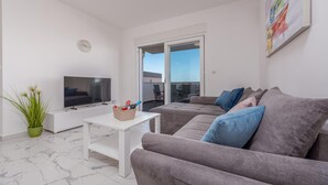 A modern living room with a flat-screen TV, a grey sofa with blue cushions, a white coffee table, and a potted plant.