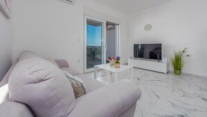 A modern living room with a grey sofa, a flat-screen TV, a small white table with decorative items, and a potted plant. - 2 bedroom amazing apartment in Povljana (Povljana)