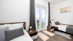 A modern hotel room with two beds, a large window with a view, a small table with a lamp, and a painting on the wall.