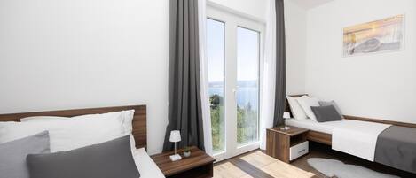 A modern hotel room with two beds, a large window with a view, a small table with a lamp, and a painting on the wall.